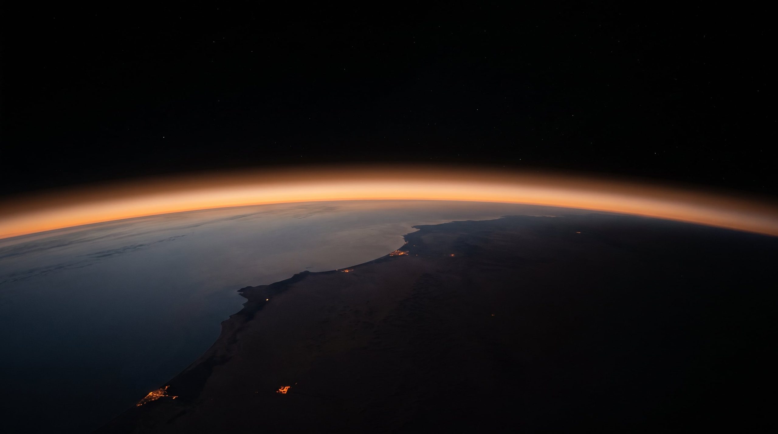 The curve of the Earth from orbit at late sunset: a desert coastline under a thin orange band of atmosphere, a few city lights on the shore, black space above.